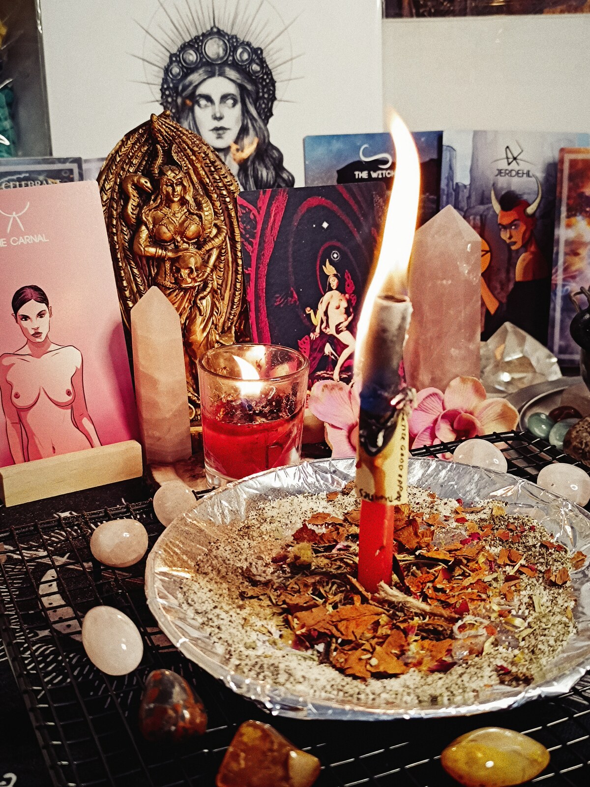 A burning Hot Girl X Appeal petition candle surrounded by herbs, rose quartz, flowers, cards, and Lilith imagery
