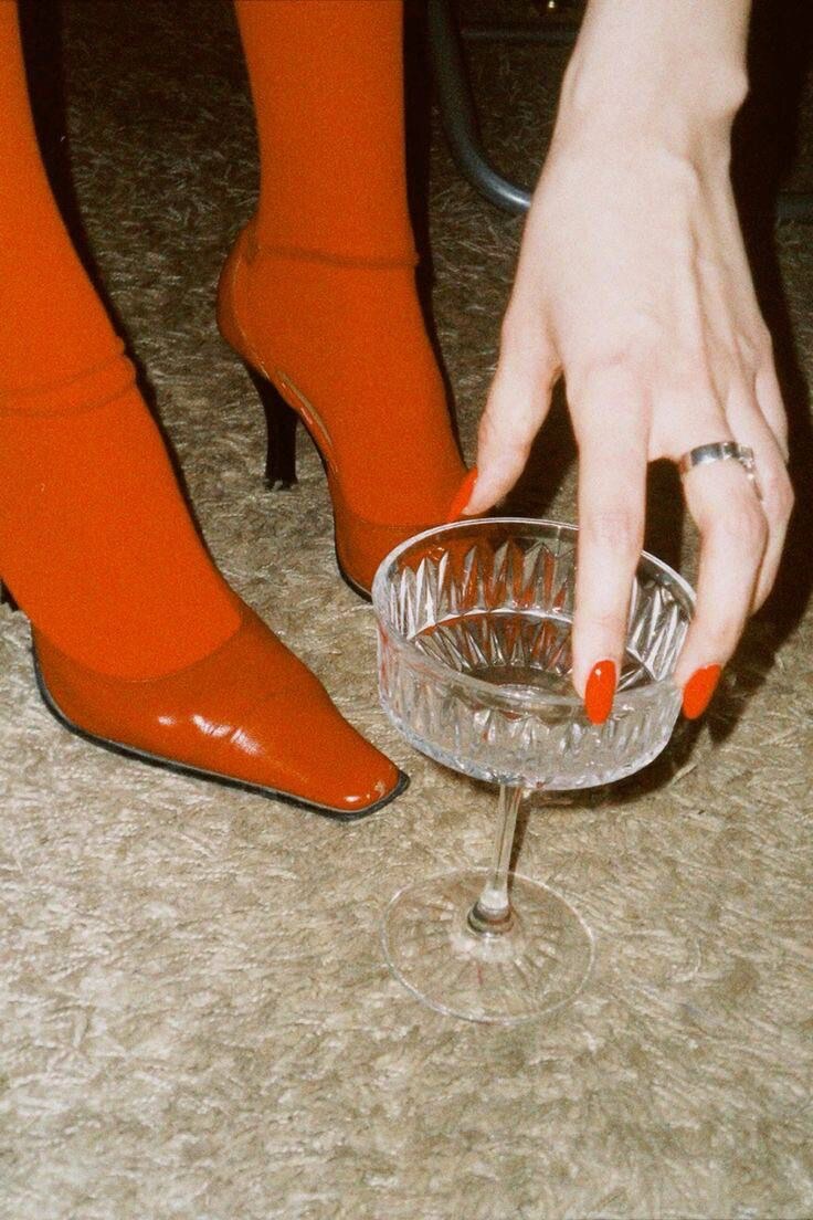 A red-manicured hand reaching toward a crystal glass beside vivid red boots, suggesting desire held with intention