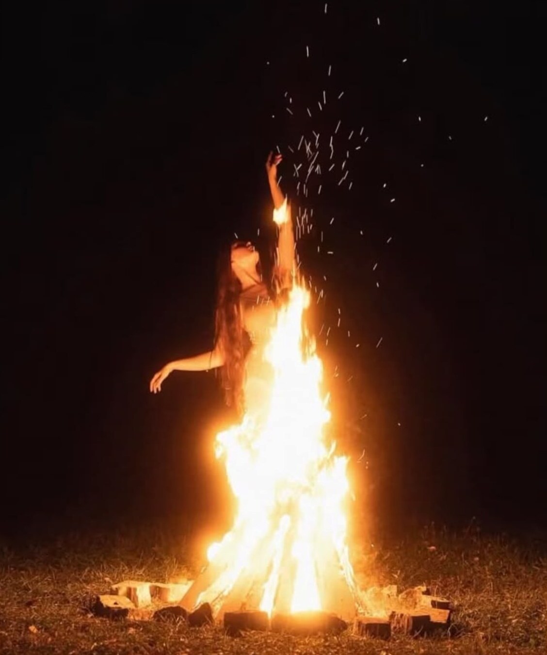 A figure standing with one arm raised beyond a bright ritual fire at night