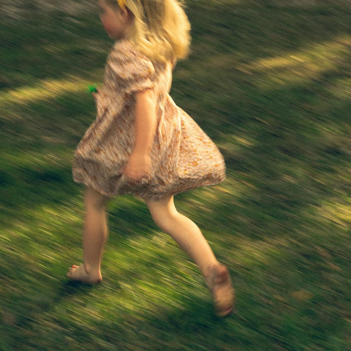 A barefoot child runs across green grass in a light patterned dress